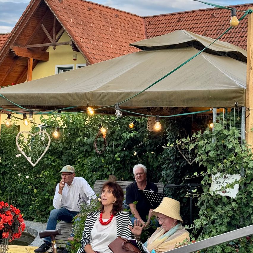 Drei Menschen sitzen unter einem Zelt im Garten. Eine Frau liest ein Buch, während die beiden Männer zuhören. Der Garten ist mit roten Blumen, Lichterketten und einem Herz geschmückt.