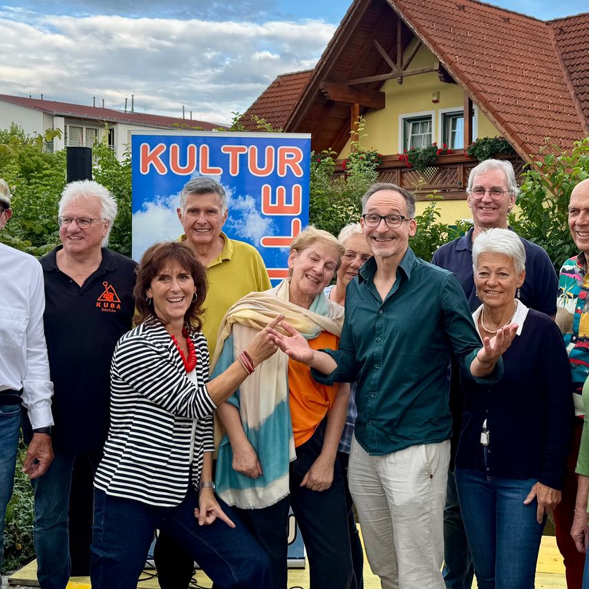 Eine Gruppe von Erwachsenen, sowohl Männer als auch Frauen, steht gemeinsam für ein Foto vor einem Haus mit einem Banner, das in großen Buchstaben 'Kultur' steht.