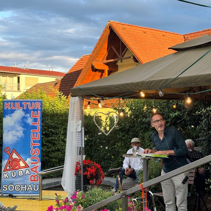 Ein Mann spricht in ein Mikrofon auf einer Bühne mit einem Zelt über ihm, in der Nähe eines Schildes, das Kultur Baustelle Kuba Sochau sagt. Hinter ihm sitzt ein anderer Mann an einem Tisch mit einem Glas, und eine dritte Person ruht auf einer Bank.