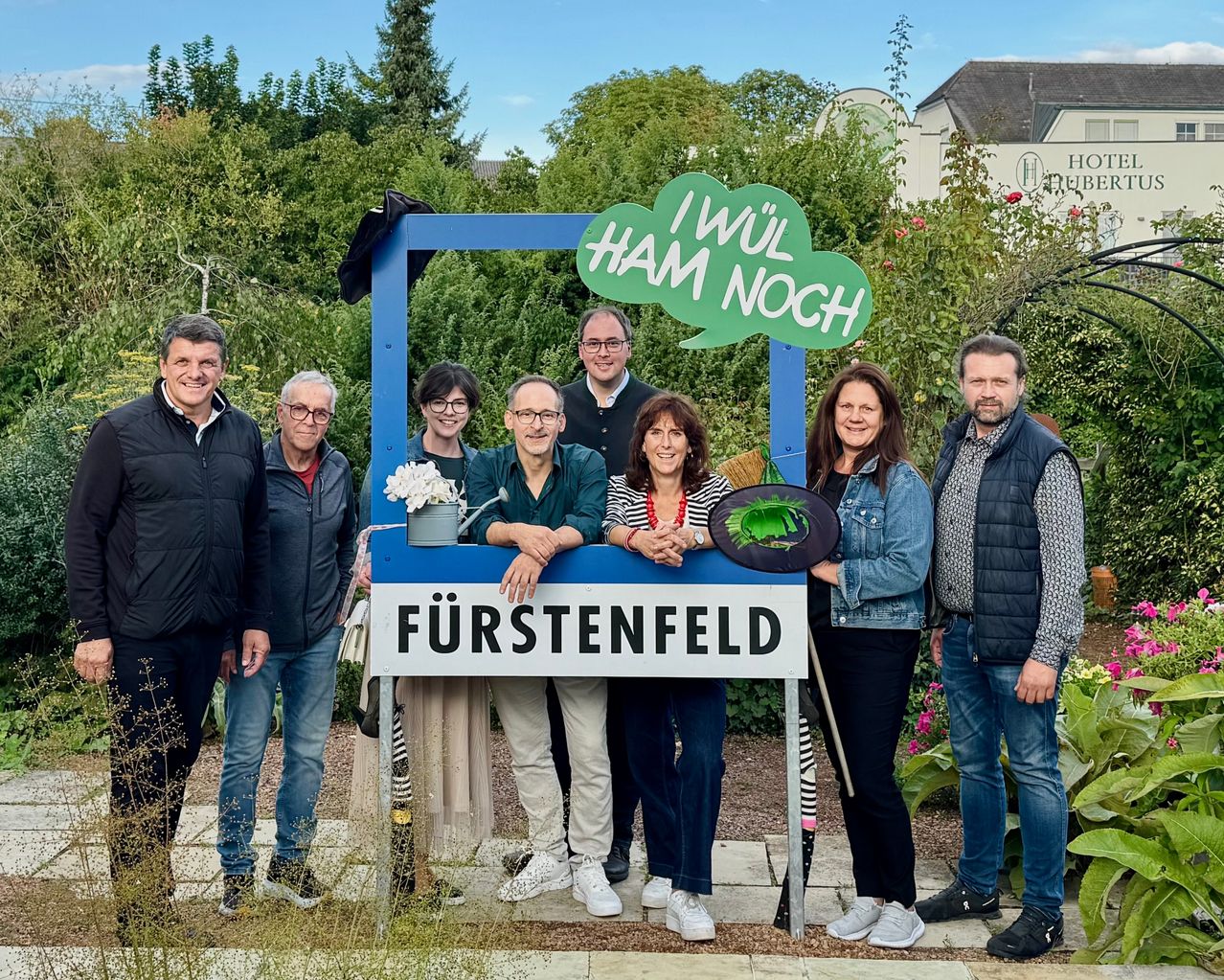 Eine Gruppe von Menschen posiert für ein Foto vor einem Stand mit dem Text 'Fürstenfeld' und einer grünen Sprechblase mit dem Text 'Ich will noch ham' in einem Garten.