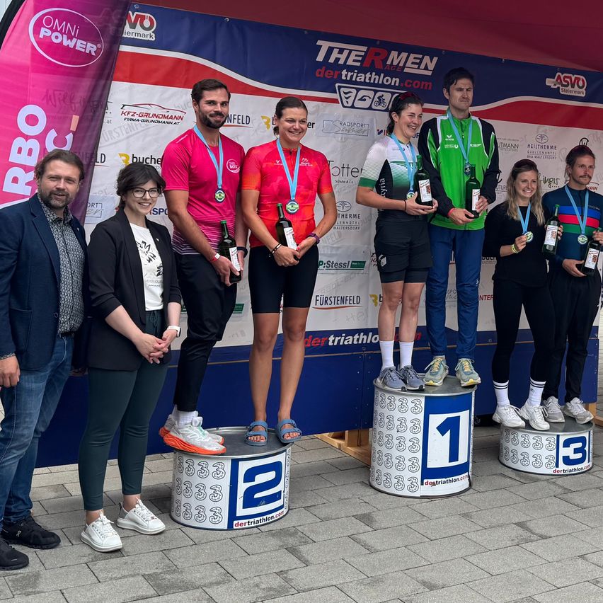 Sieger eines Triathlon-Events stehen auf einem Podium mit Medaillen um den Hals. Der Erstplatzierte hält eine Flasche Wein in der Hand.