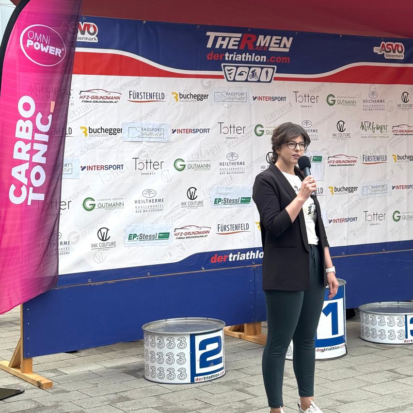 Eine Frau in einem schwarzen Blazer und grünen Hosen spricht in ein Mikrofon, stehend vor einem Plakat, das für The R Men Triathlon wirbt. Ein rosa Banner mit den Worten 'CARBO TONIC' und ein Fass, das mit '2' markiert ist, sind in der Nähe.