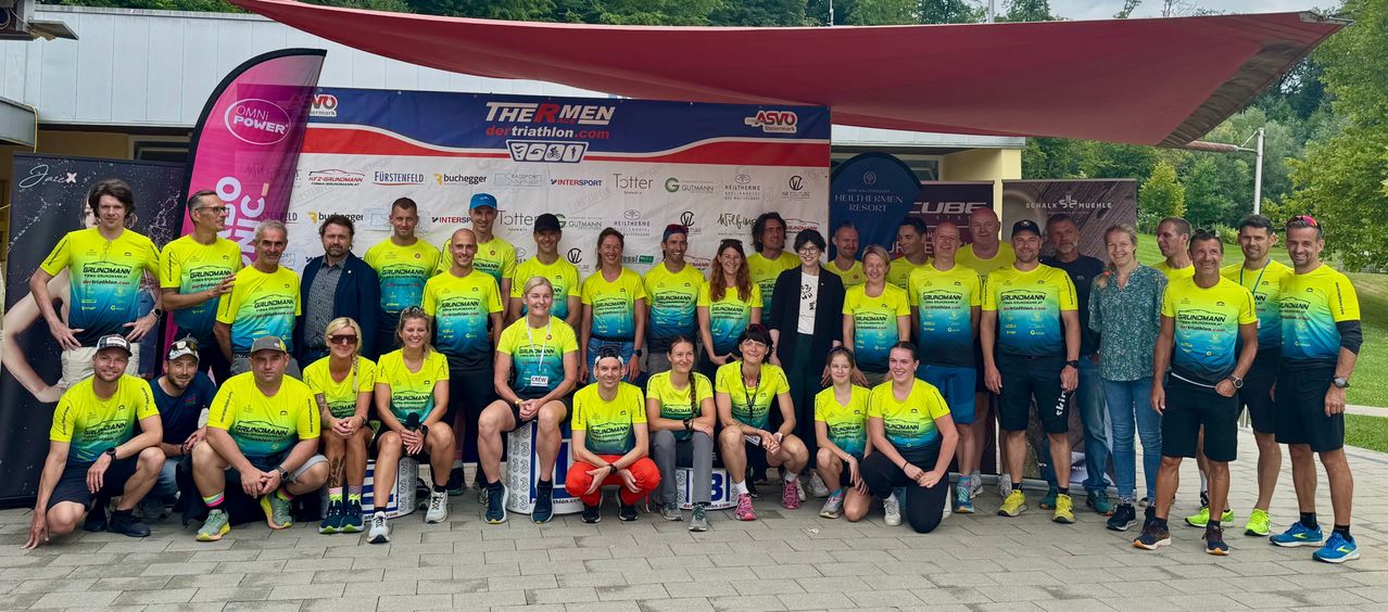 Eine Gruppe von Menschen in gelben Shirts posiert für ein Foto vor einem Banner, das 'The RMEN dertriathlon.com' besagt. Sie lächeln und einige tragen eine Brille. Dahinter sind weitere Menschen und ein Gebäude mit einem Banner. In der Ferne sind auch einige Bäume zu sehen.