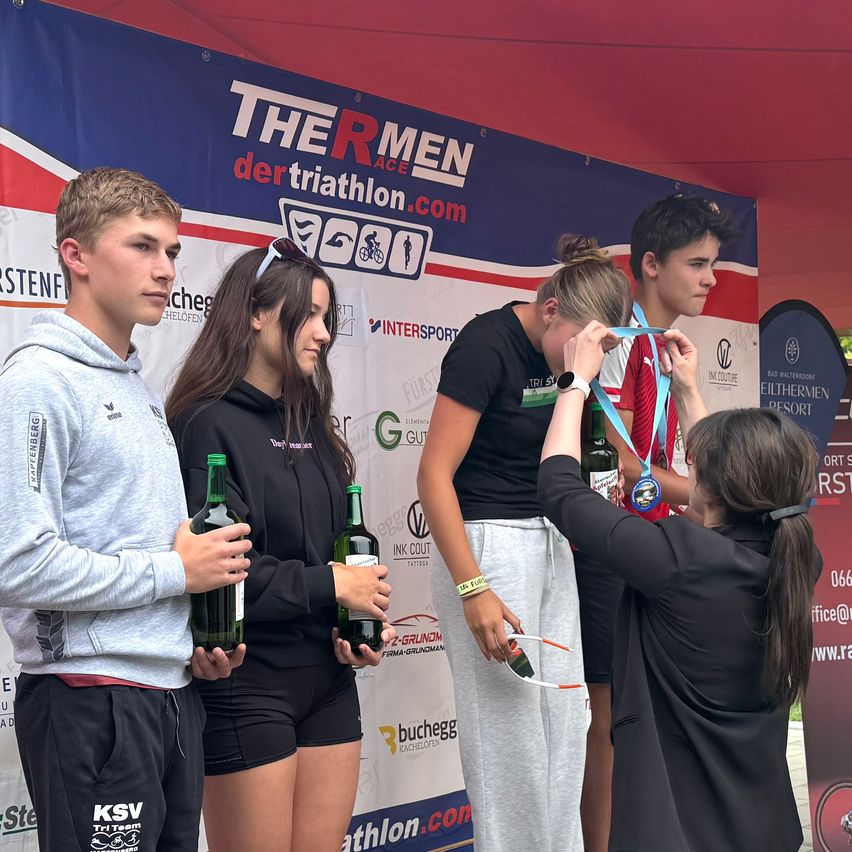 Eine Gruppe von Athleten steht auf einem Podium und hält Flaschen. Die Frau in der Mitte erhält eine Medaille von einer anderen Frau. Hinter ihnen befindet sich ein Banner, das für 'The R Men der Triathlon.com' wirbt.