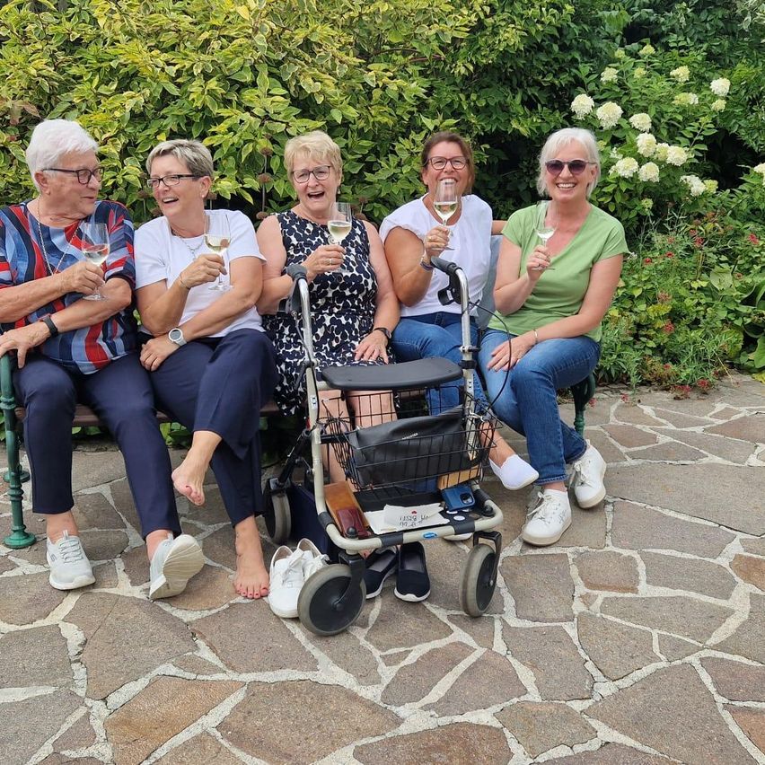 Fünf Frauen sitzen auf einer Bank und halten Weingläser in der Hand. Sie lächeln und scheinen für ein Foto zu posieren. Die Frau auf der rechten Seite benutzt einen Rollator.