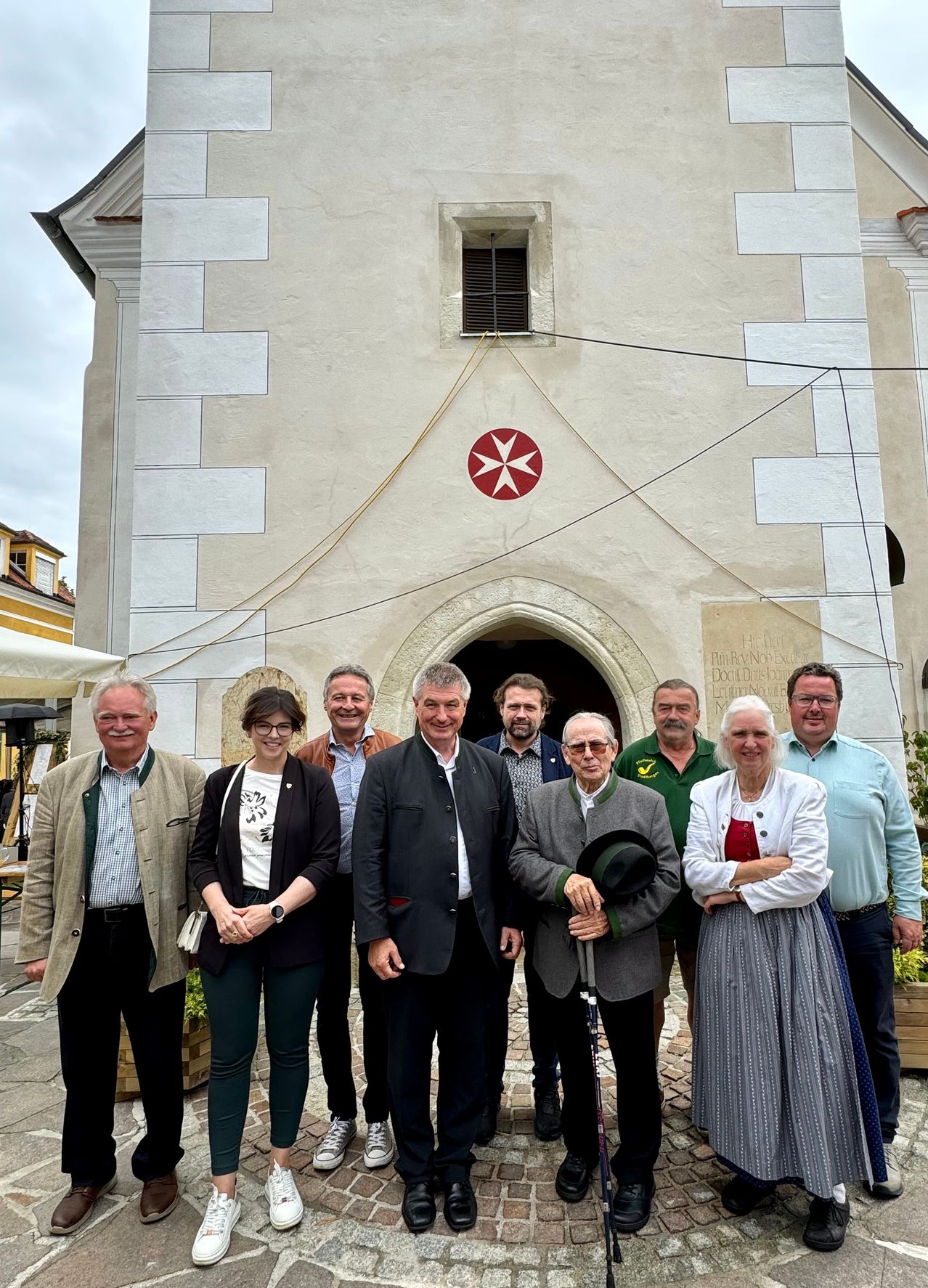 Eine Gruppe von Menschen steht vor einer Kirche, lächelt und posiert für ein Foto. Die Kirche hat ein rotes maltesisches Kreuzemblem an ihrer Wand.
