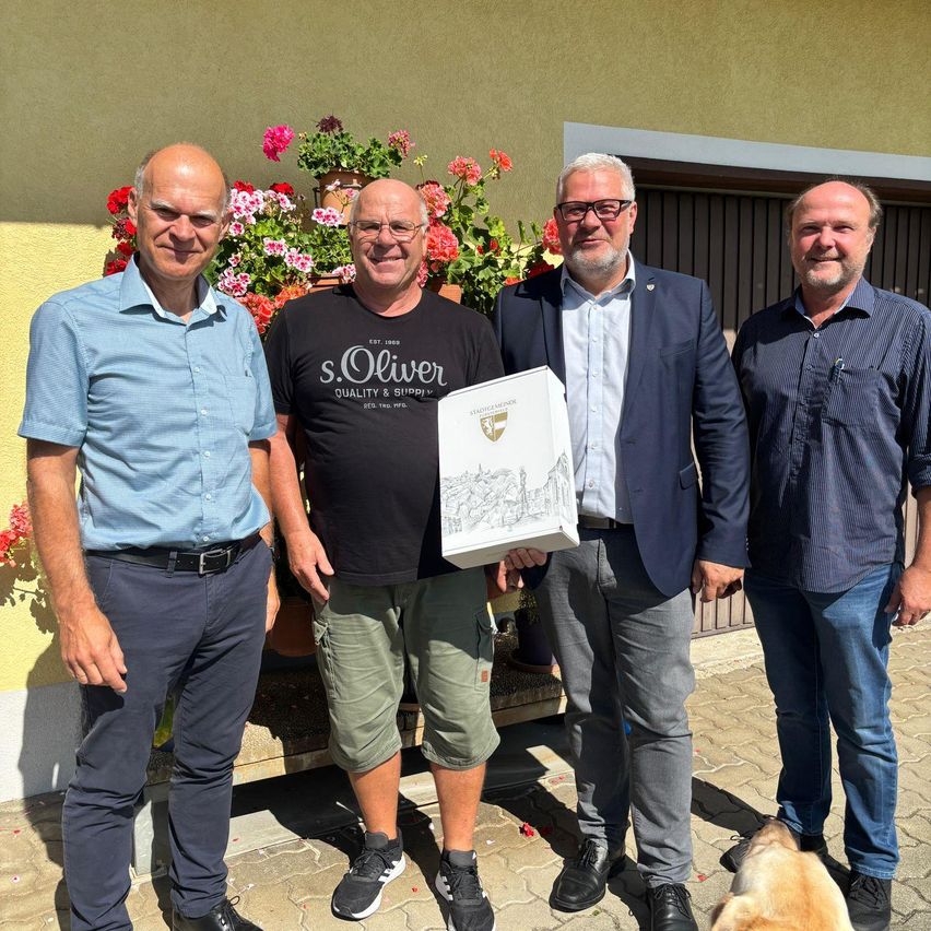 Vier Männer stehen vor einer gelben Wand mit bunten Blumen und einem Hund. Der Mann in der Mitte hält ein Zertifikat, und der Mann rechts trägt eine Brille.