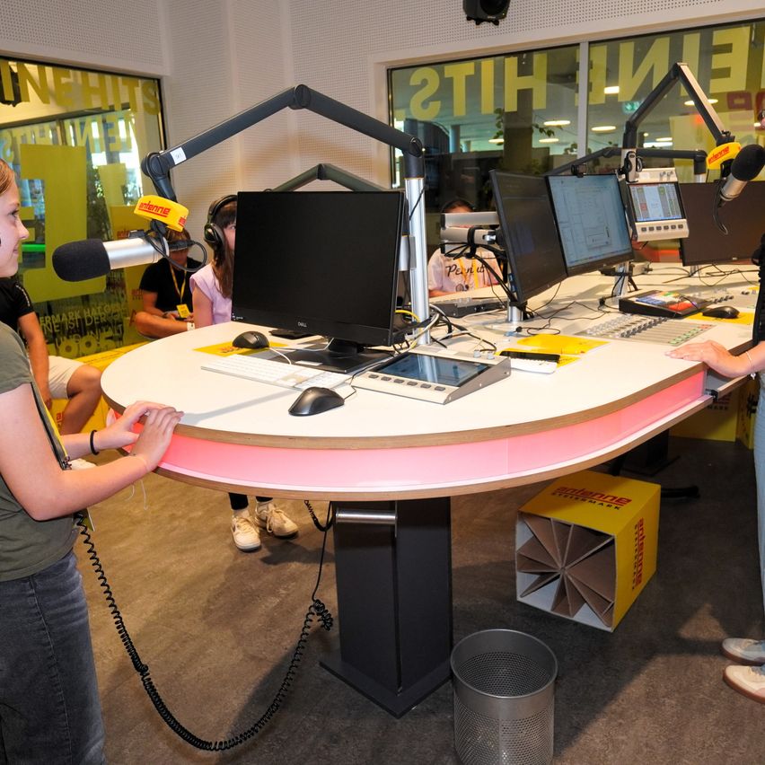 Ein Radiostudio mit zwei Personen, die die Ausrüstung bedienen, eine hält ein Mikrofon. Ein Schreibtisch mit Monitoren, Tastaturen und anderen Büromaterialien. Ein Mülleimer ist in der Nähe.