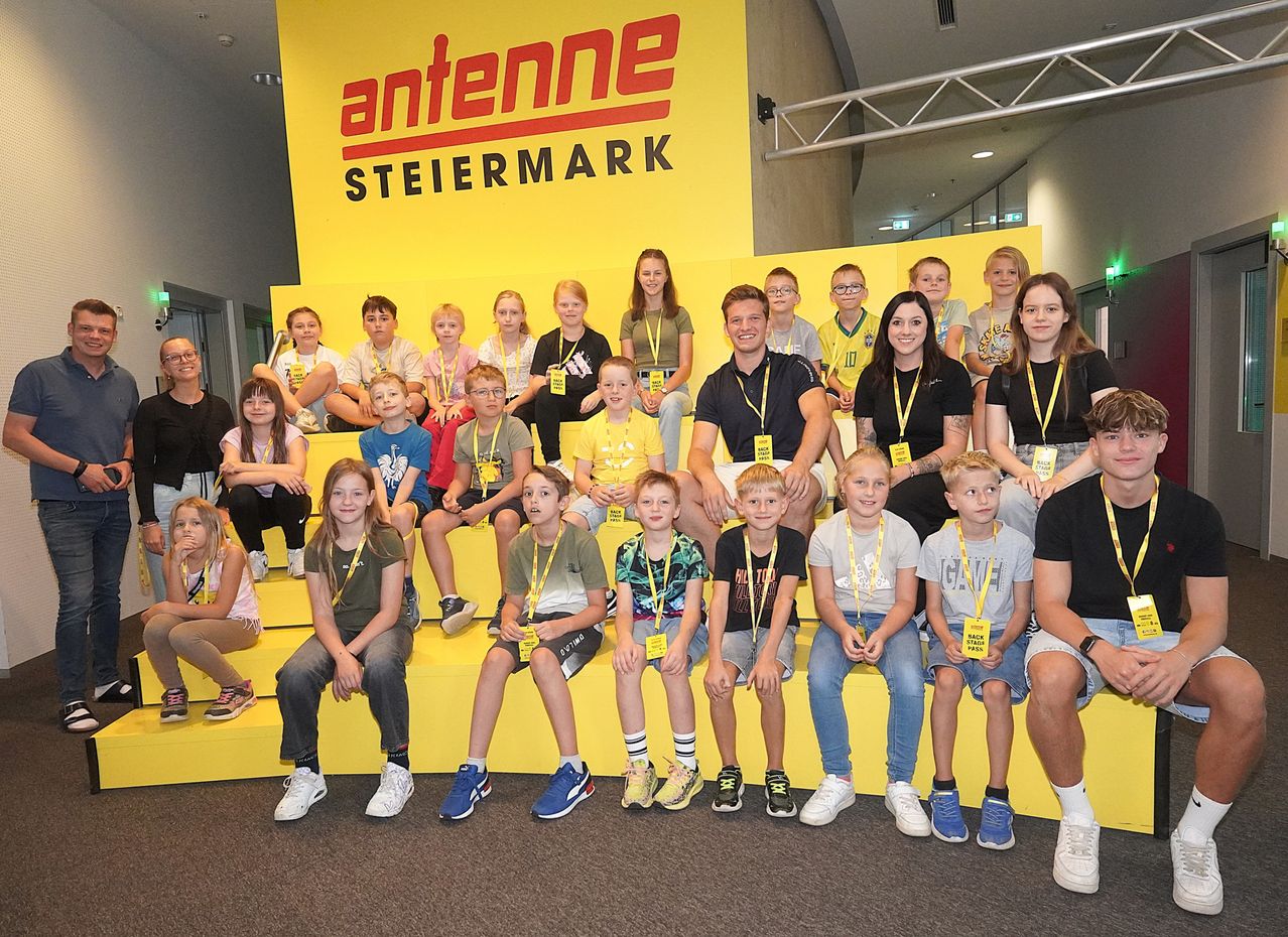 Eine Gruppe von Kindern und Erwachsenen posiert für ein Foto vor einem gelben Antenna Steiermark-Schild. Einige tragen gelbe Lanyards, andere verschiedene farbige Kleidung. Sie sitzen auf gelben Bänken.
