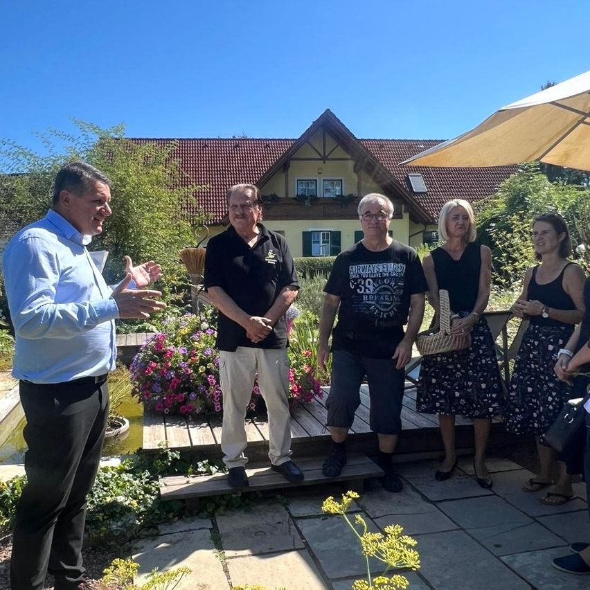 Eine Gruppe von Menschen steht in einem Garten mit einem gelben Haus im Hintergrund. Ein Mann in einem blauen Hemd spricht, während andere zuhören. Der Garten hat bunte Blumen und Pflanzen.
