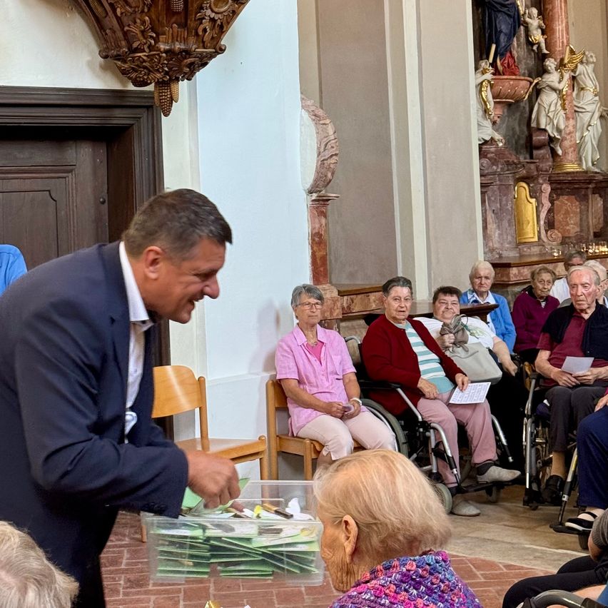 In einer Kirche verteilt ein Mann Umschläge an sitzende Senioren. Hinter ihm schmücken zwei Statuen die Wand. Senioren sitzen auf Stühlen, einer im Rollstuhl, und einige halten Papiere in den Händen.