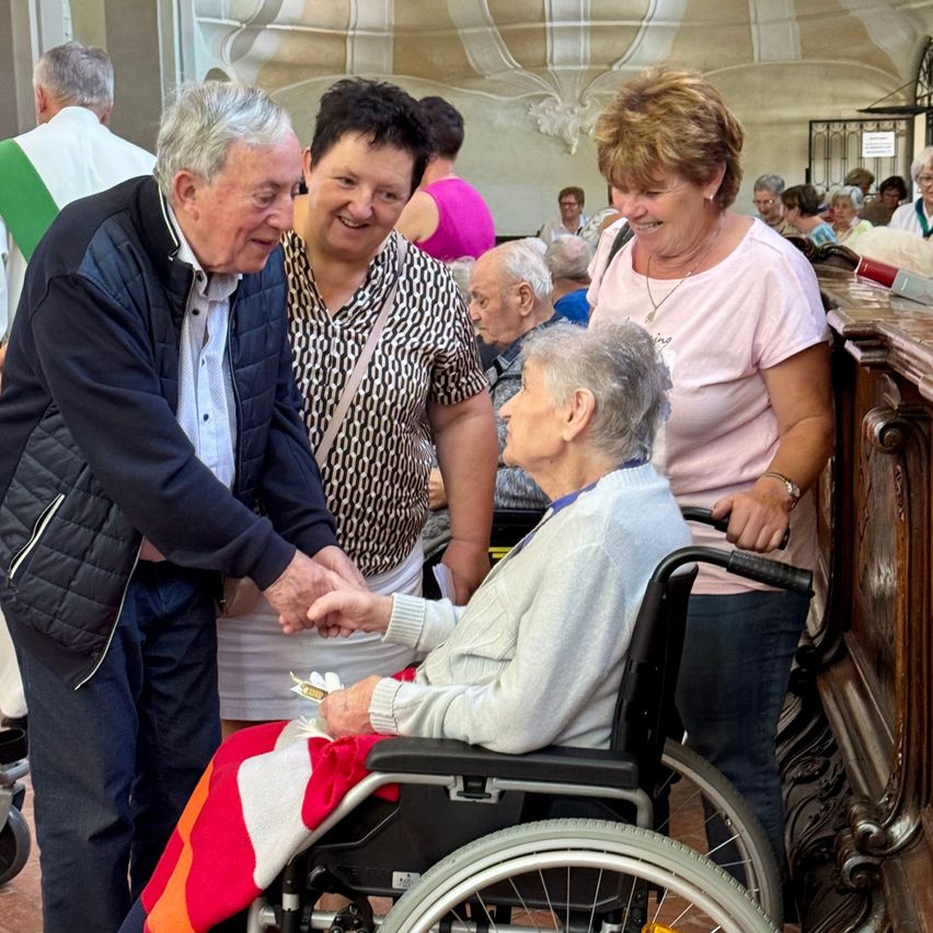 Eine Gruppe älterer Menschen versammelt sich in einer Kirche. Ein älterer Mann schüttelt einer Frau im Rollstuhl die Hand, während andere zusehen.