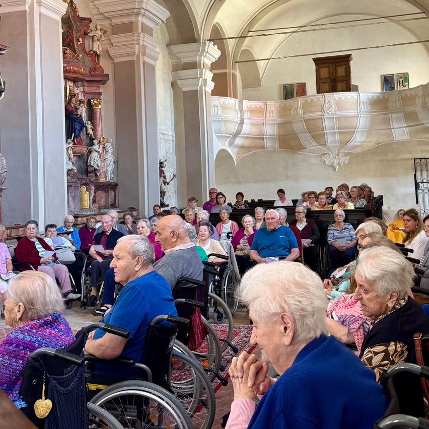 Eine Gruppe älterer Menschen in einer Kirche, einige in Rollstühlen, lauschen aufmerksam. Im Hintergrund befindet sich eine Statue und ein Altar.
