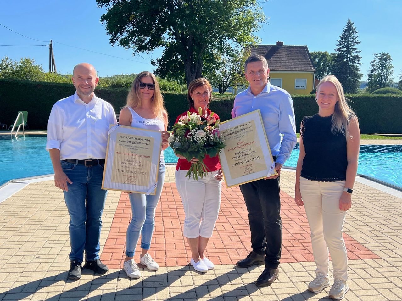 Fünf Personen stehen vor einem Pool, halten Urkunden und Blumen. Sie lächeln und scheinen eine Errungenschaft zu feiern. Der Mann links trägt einen Gürtel, und die Frau in der Mitte hält einen Blumenstrauß. Im Hintergrund befindet sich ein Haus mit einem Baum davor.