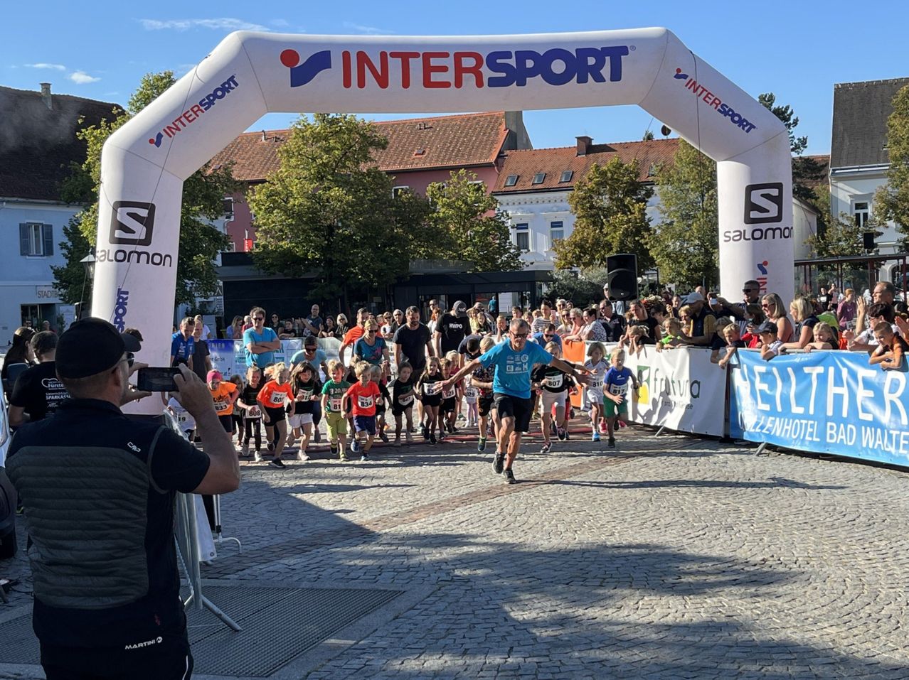 Ein Marathon ist im Gange, wobei die Läufer unter einem INTERSPORT-Bogen hindurchlaufen. Zuschauer beobachten von hinter einem Banner aus. Bäume und Gebäude sind im Hintergrund.