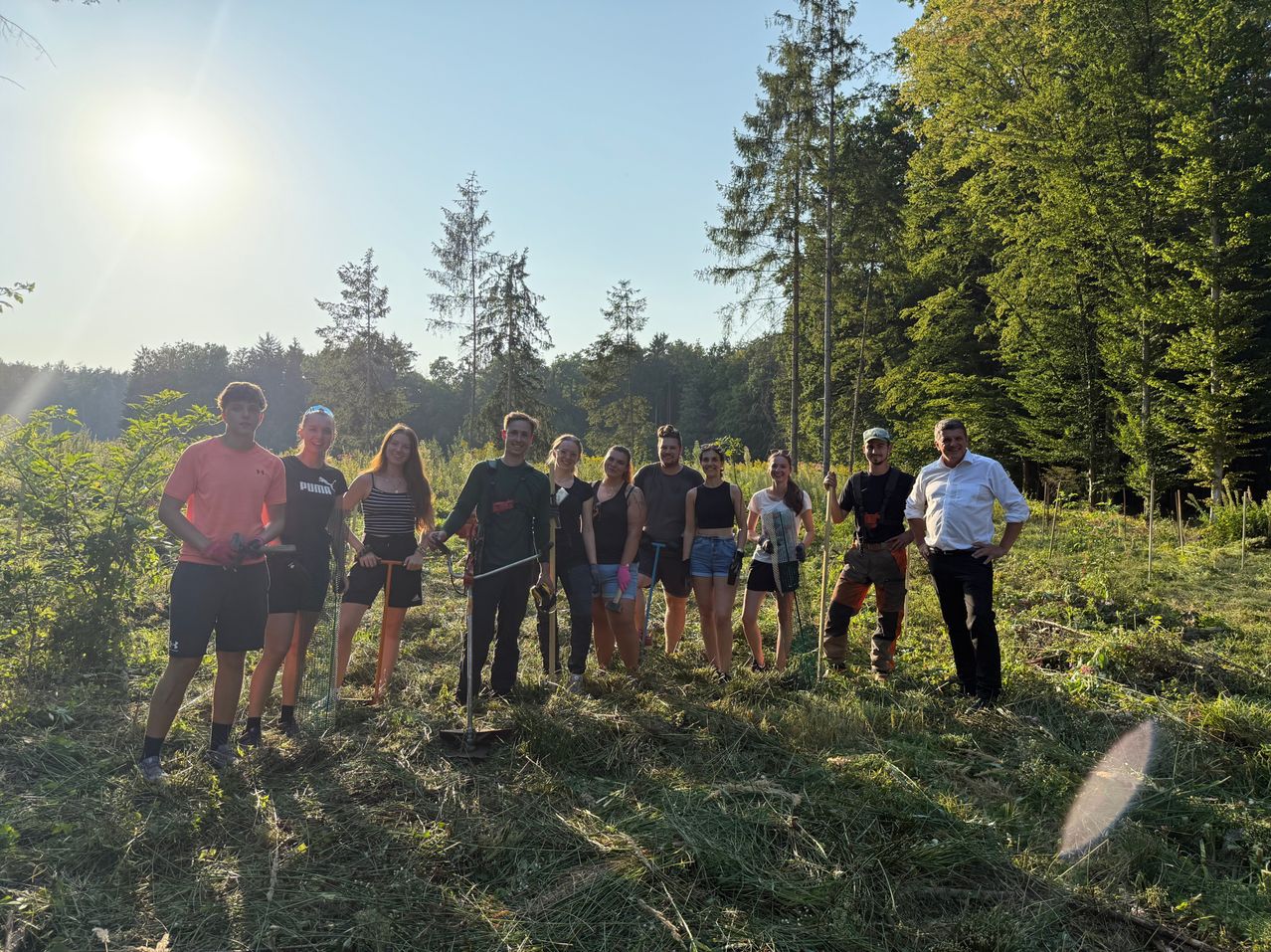 Eine Gruppe von Menschen, wahrscheinlich Freiwillige, posiert für ein Foto in einem Grasfeld mit Bäumen im Hintergrund. Sie halten Gartengeräte in der Hand und tragen Freizeitkleidung.