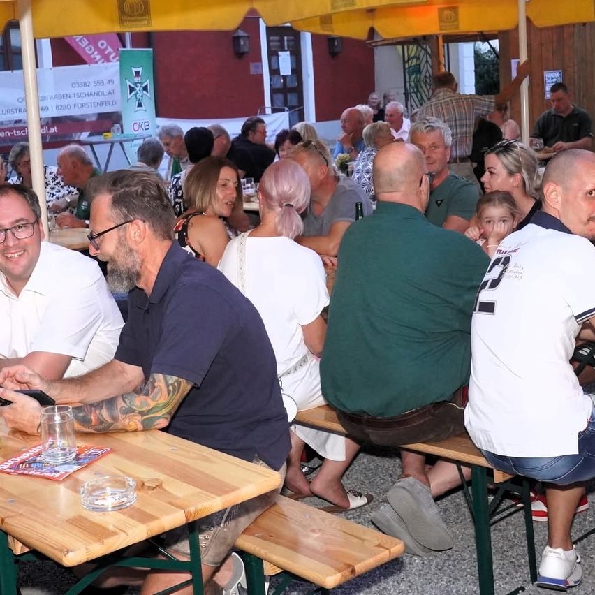 Eine Gruppe von Menschen sitzt unter einem gelben Regenschirm an einem Tisch. Sie scheinen in ein Gespräch vertieft zu sein. Ein Banner ist im Hintergrund sichtbar.