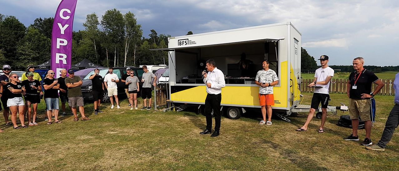 Ein Mann hält eine Rede mit einem Mikrofon vor einem mobilen Sicherheitsstand, andere stehen und lauschen, mit geparkten Autos und Bäumen im Hintergrund.