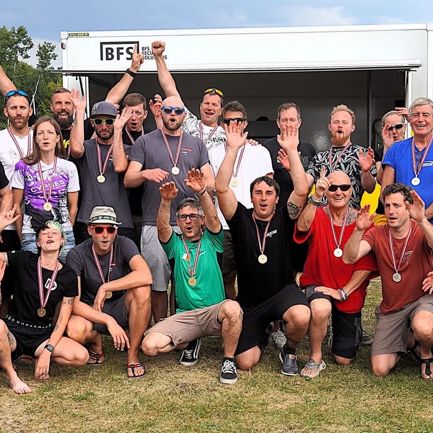 Eine Gruppe von Männern und Frauen mit Medaillen posiert für ein Foto im Freien. Sie stehen und sitzen auf Gras, mit einem weißen Lastwagen mit der Aufschrift 'BFS' im Hintergrund.