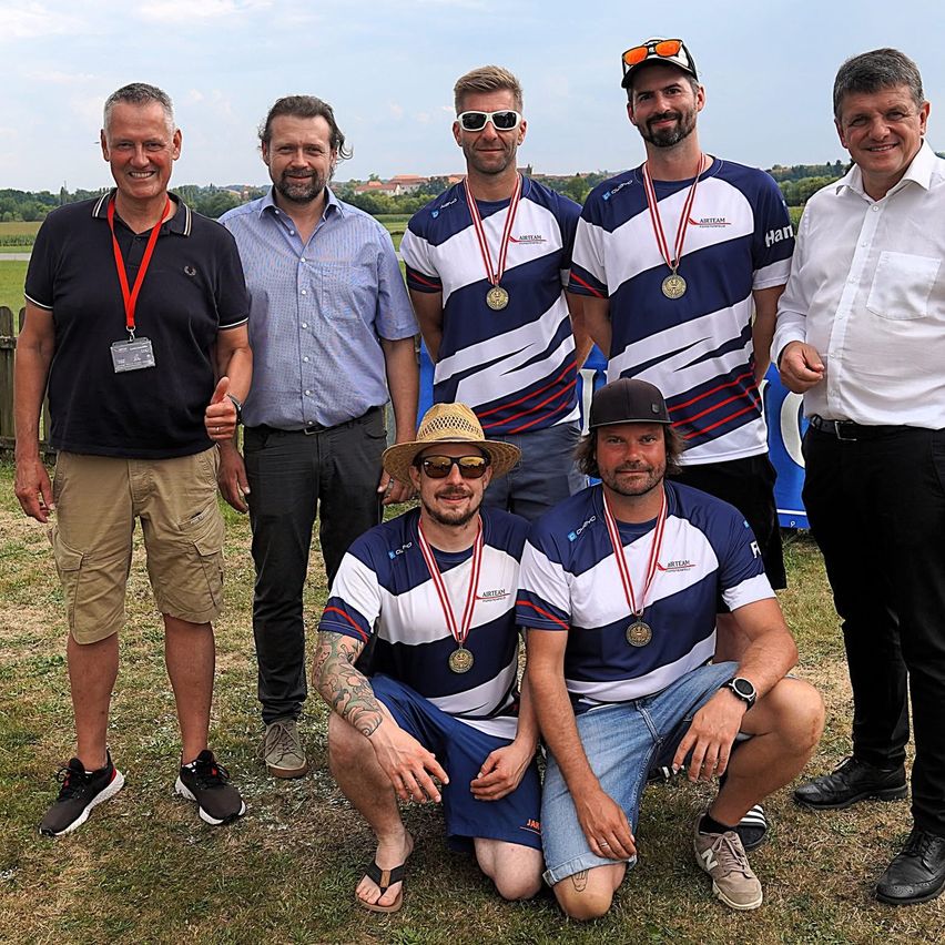 Eine Gruppe von Männern mit Medaillen und Freizeitkleidung posiert für ein Foto auf einem Grasfeld, einer kniet. Hinter ihnen ist im Hintergrund ein Zaun und Bäume zu sehen.