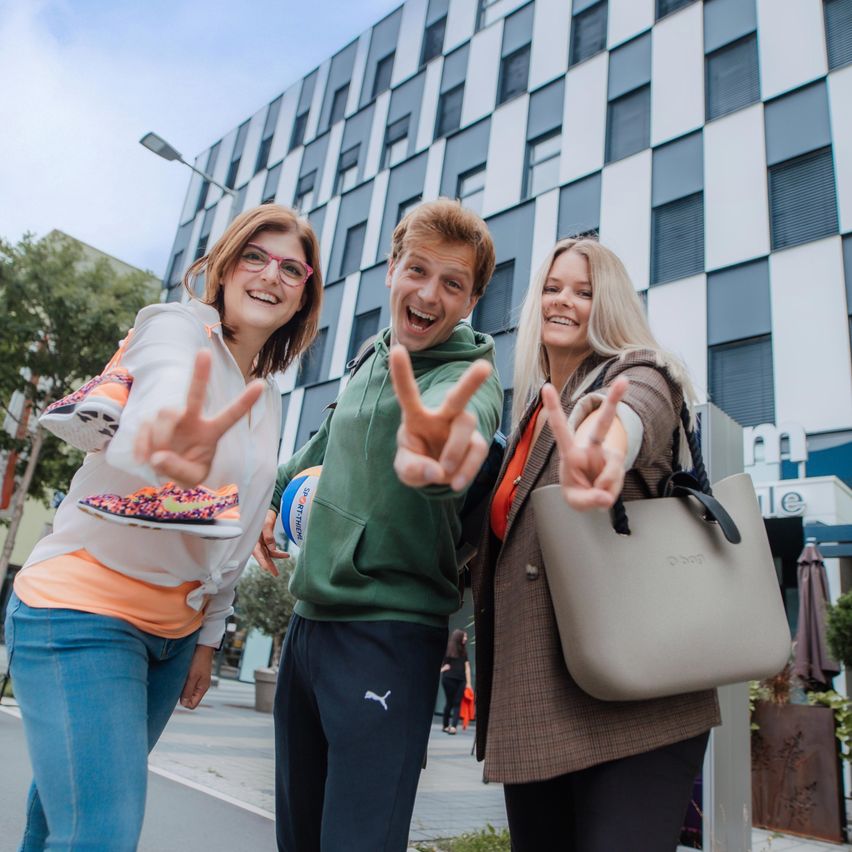 Drei Personen stehen vor einem Gebäude, zwei machen Friedenszeichen, eine hält einen Sportball. Sie lächeln und scheinen für ein Foto zu posieren.