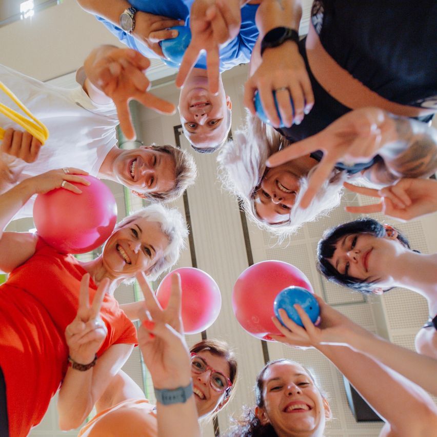 Eine Gruppe von Menschen steht im Kreis mit rosa und blauen Bällen, lächelt und macht Friedenszeichen. Der Raum hat eine weiße Decke und weiße Wände.