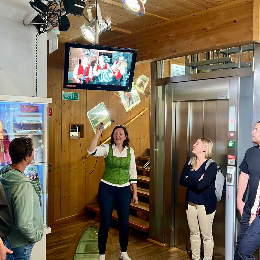In einem Raum hält eine Frau eine Taschenlampe und zeigt mit ihr nach oben zu einem hängenden Fernseher. Daneben steht eine weitere Frau, die zuschaut. In der Nähe stehen zwei Männer neben einem Aufzug. Die Wände sind aus Holz, mit einem Kühlschrank und verschiedenen Schildern im Hintergrund.
