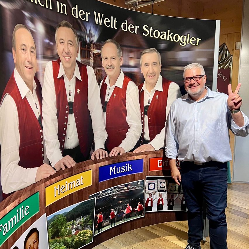 Ein Mann steht vor einem Banner mit vier lächelnden Männern in traditioneller Kleidung. Sie haben ihre Hände auf einem Banner mit verschiedenen Kategorien. Der Mann trägt eine Brille und eine Uhr.