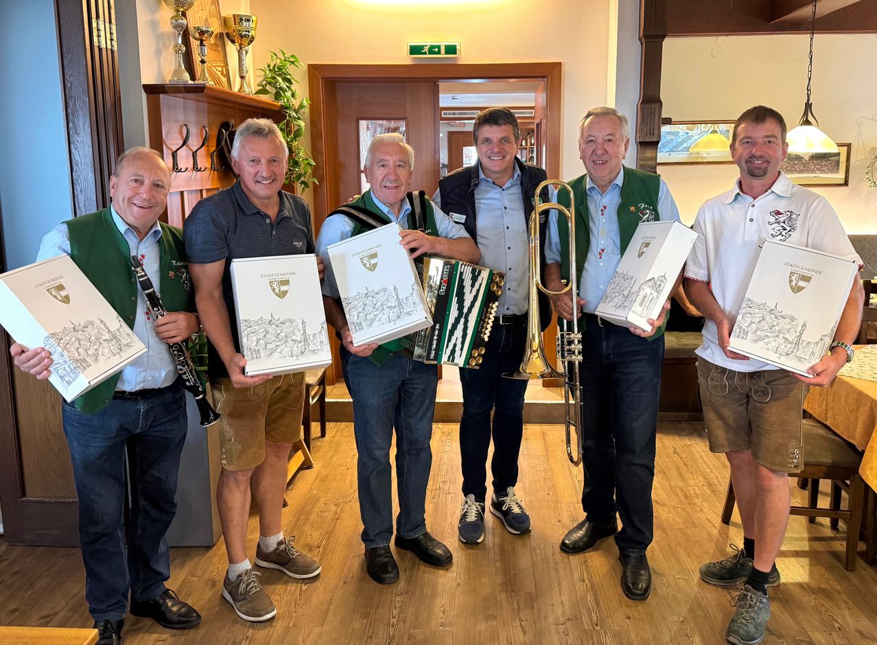 Fünf Männer posieren drinnen und halten Musikinstrumente und Bücher. Ein Mann hält eine Trompete, ein anderer ein Akkordeon. Hinter ihnen eine Holztür und ein Regal mit Trophäen.