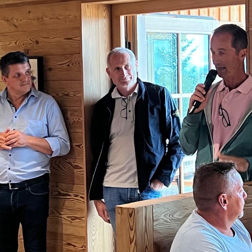 Vier Männer stehen in einem Raum mit Holzwänden, einer spricht in ein Mikrofon. Der Mann auf der linken Seite trägt ein blaues Hemd und Jeans. Der Mann neben ihm trägt eine schwarze Jacke und eine Brille. Der dritte Mann trägt ein rosa Hemd und spricht in ein Mikrofon. Der Mann auf der rechten Seite trägt ein weißes Hemd.