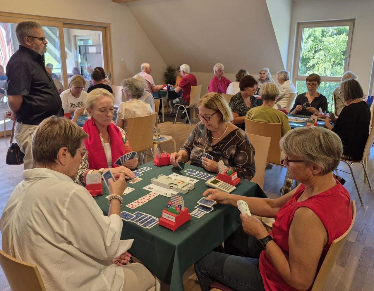 Eine Gruppe älterer Frauen spielt Kartenspiele an einem Tisch in einem Raum mit Holzböden und Glasfenstern.