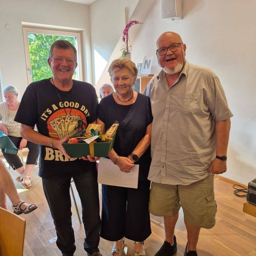 Drei Personen stehen in einem Raum, zwei Männer und eine Frau, lächeln und halten eine Geschenkbox mit Früchten und einem Papier, möglicherweise bei einer Veranstaltung.