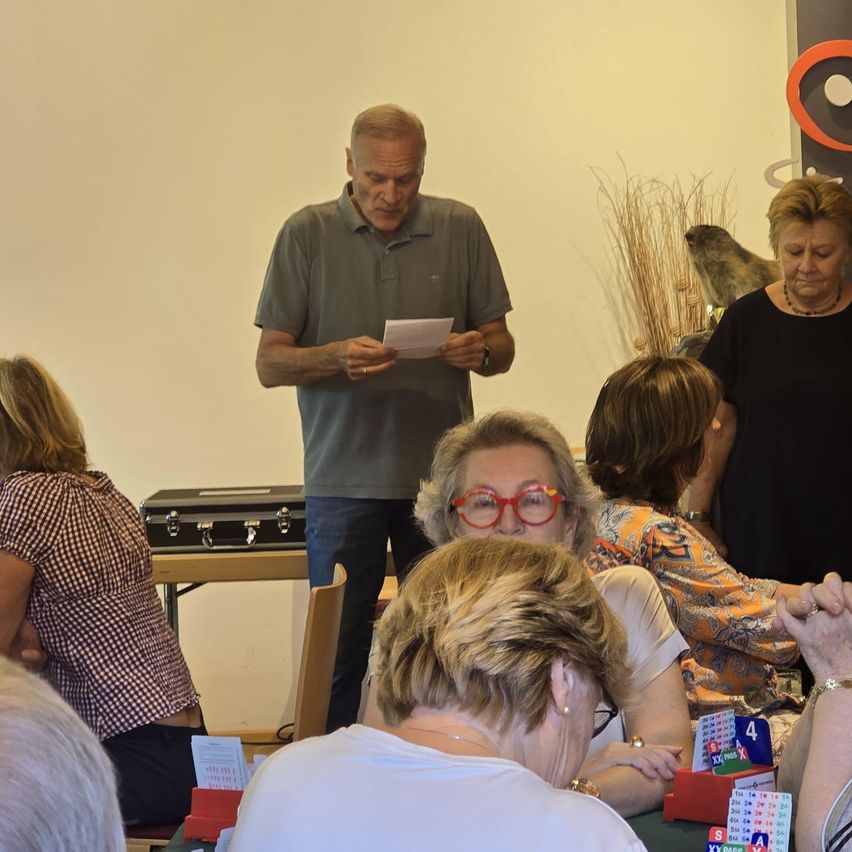 Eine Gruppe von Menschen sitzt um einen Tisch herum, einige führen Gespräche. Ein Mann liest von einem Stück Papier, während andere aufmerksam zuhören. Eine Frau trägt eine Brille, und eine Aktentasche steht auf einem nahegelegenen Tisch.