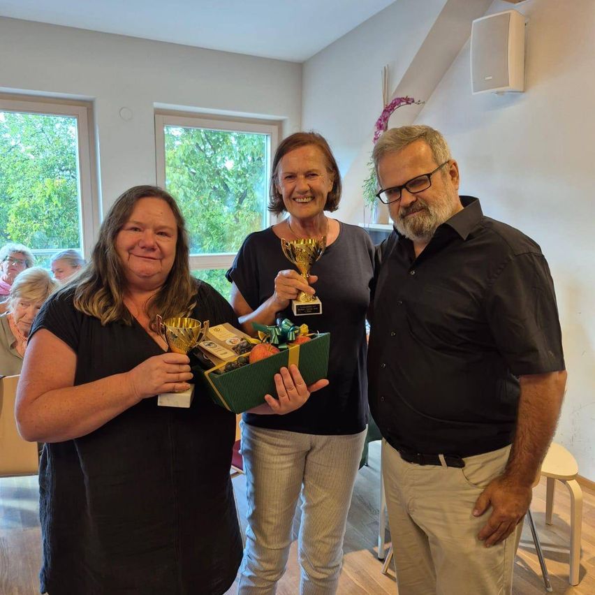 Drei Menschen stehen zusammen in einem Raum mit Geschenken und Trophäen. Zwei Frauen halten Trophäen, eine mit einer grünen Box. Ein Mann steht neben ihnen. Dahinter sitzen Menschen auf Stühlen.