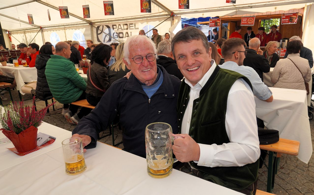Zwei Männer lächeln, halten große Gläser Bier in einer festlichen Umgebung mit Zelten und anderen Gästen.
