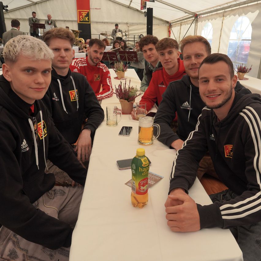 Eine Gruppe von Männern in Sporttrikots sitzt mit Getränken und Essen an einem Tisch und lächelt für ein Foto in einem Zelt mit Musikinstrumenten und Leuten im Hintergrund.