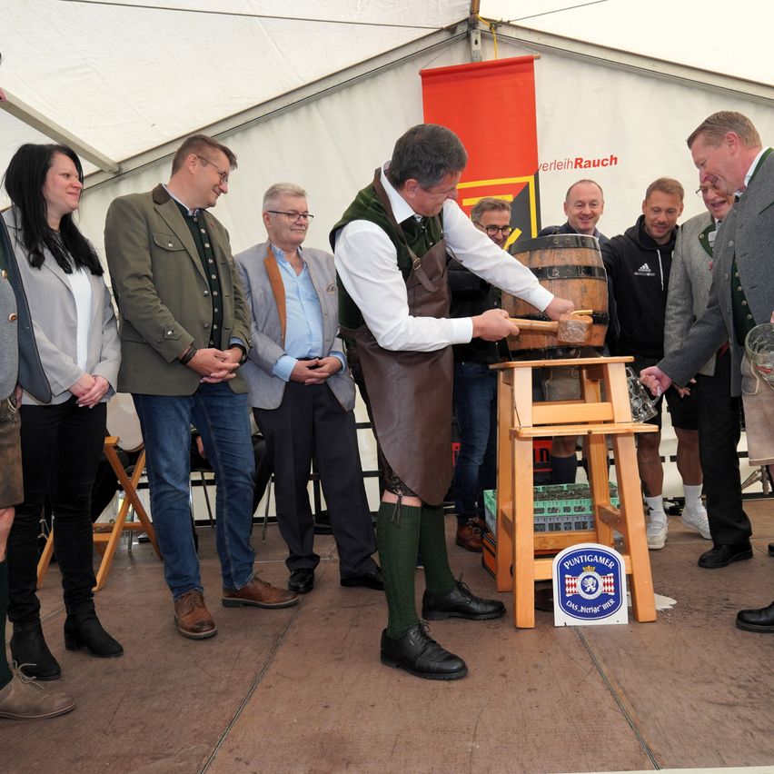 Ein Mann in traditioneller Kleidung klopft mit einem Hammer auf ein Holzfass, während andere zusehen, in einem Zelt mit einem Banner.