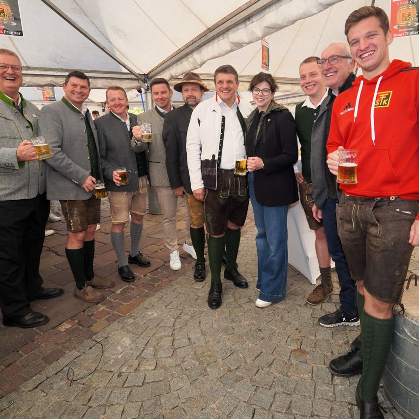 Eine Gruppe von Männern und Frauen in traditioneller deutscher Kleidung lächelt und posiert für ein Foto, während sie Biergläser halten. Sie stehen unter einem Zelt.
