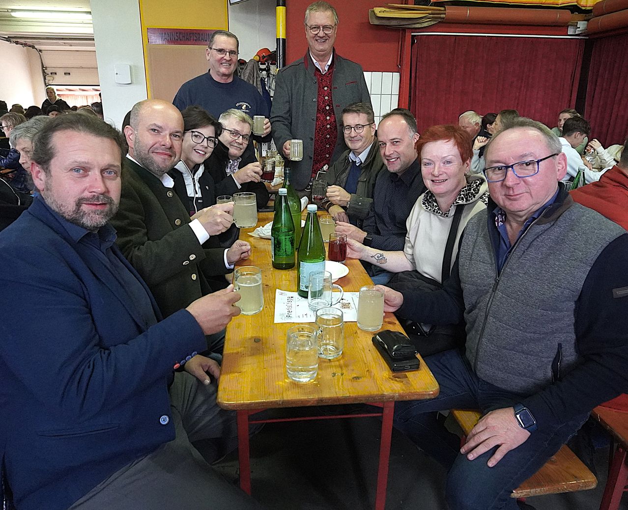 Eine Gruppe von Menschen, einige mit Brille, sitzt um einen Tisch herum, lächelt und hält Gläser mit Getränken in der Hand. Auf dem Tisch stehen Flaschen und Tassen. Dahinter steht eine Person in der Nähe einer Wand.