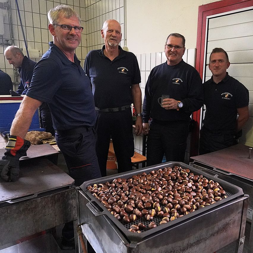 Vier Männer stehen lächelnd in einer Küche. Einer hält ein Getränk. Tabletts mit Kastanien sind auf einem Grill. Eine Tür und geflieste Wände sind im Hintergrund.