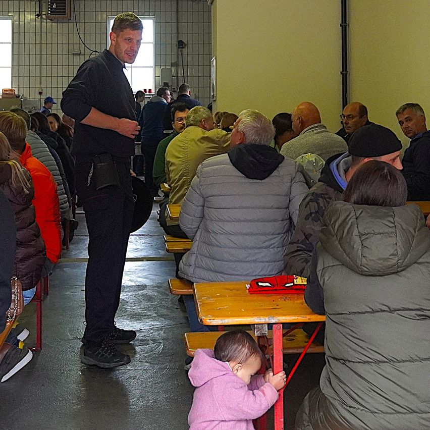 Ein Mann steht in einem überfüllten Raum, andere sitzen an Tischen. Eine Frau mit einem Baby an einem Tisch schaut den Mann an.