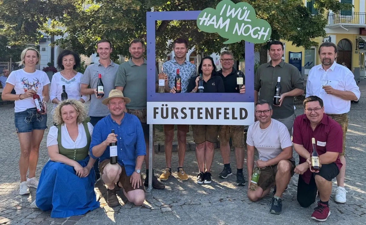 Eine Gruppe von Menschen steht vor einem Rahmen mit den Worten 'Fürstenfeld' und einem Slogan auf Deutsch. Sie lächeln und halten Weinflaschen. Einige sind in traditioneller Kleidung.