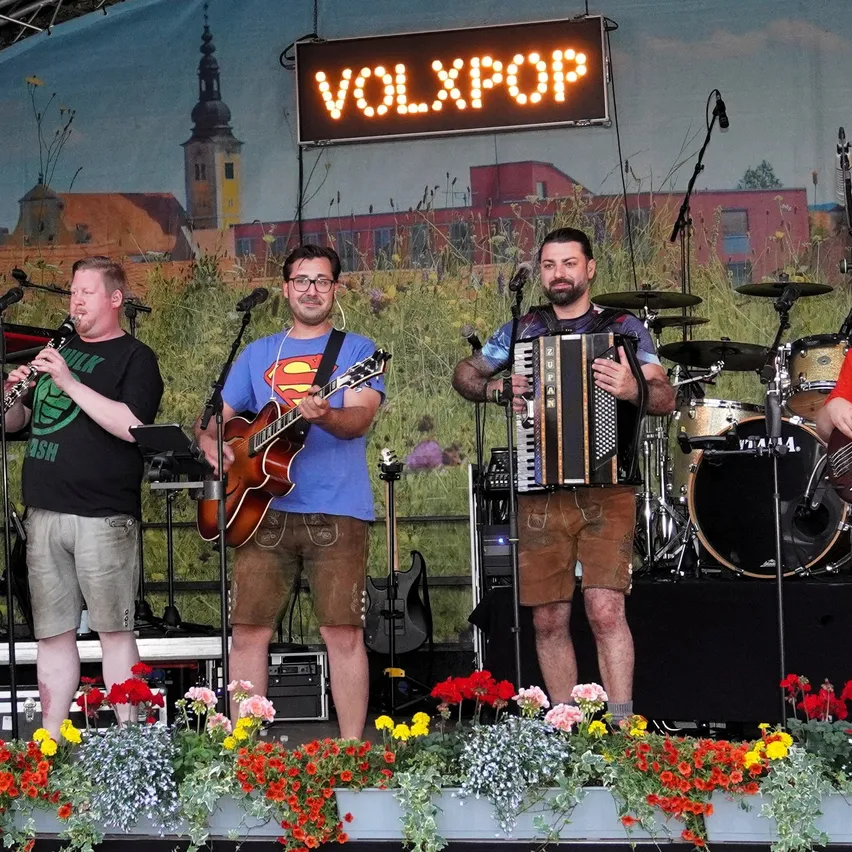 Drei Musiker treten mit einer Stadtkulisse auf der Bühne auf. Einer spielt Klarinette, ein anderer Gitarre und der dritte Akkordeon. Dahinter stehen Trommeln und andere Instrumente.