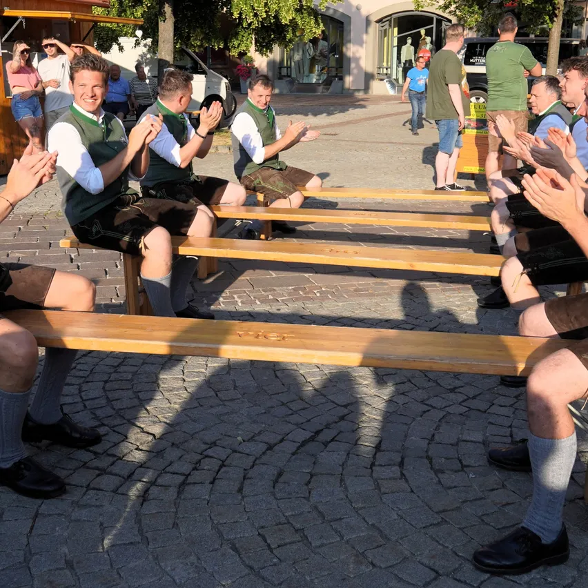 Eine Gruppe von Männern sitzt auf Holzbänken im Freien, gekleidet in traditionelle Kleidung, klatscht und lächelt. Hinter ihnen stehen oder sitzen mehrere Personen, und ein Auto ist in der Nähe geparkt.