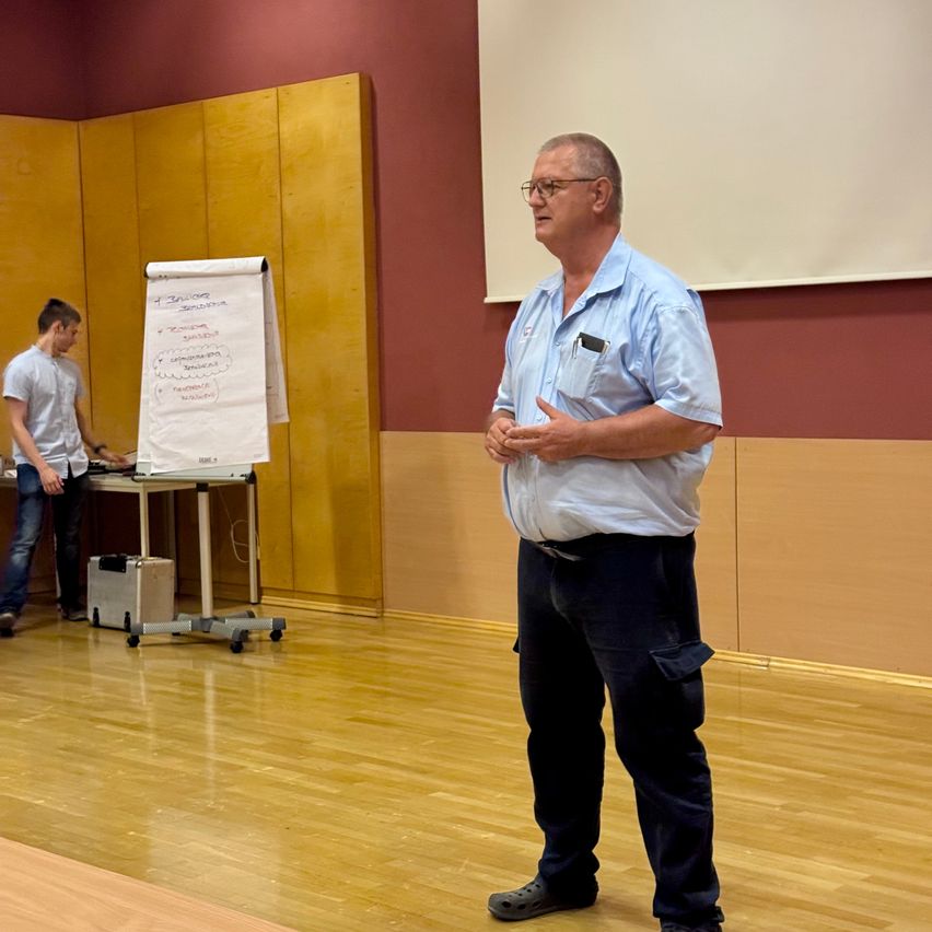 Ein Mann steht und spricht in einem Raum. Ein anderer Mann steht hinter ihm und schaut auf eine Tafel auf einem Tisch. Der Raum hat Holzböden und Wände.