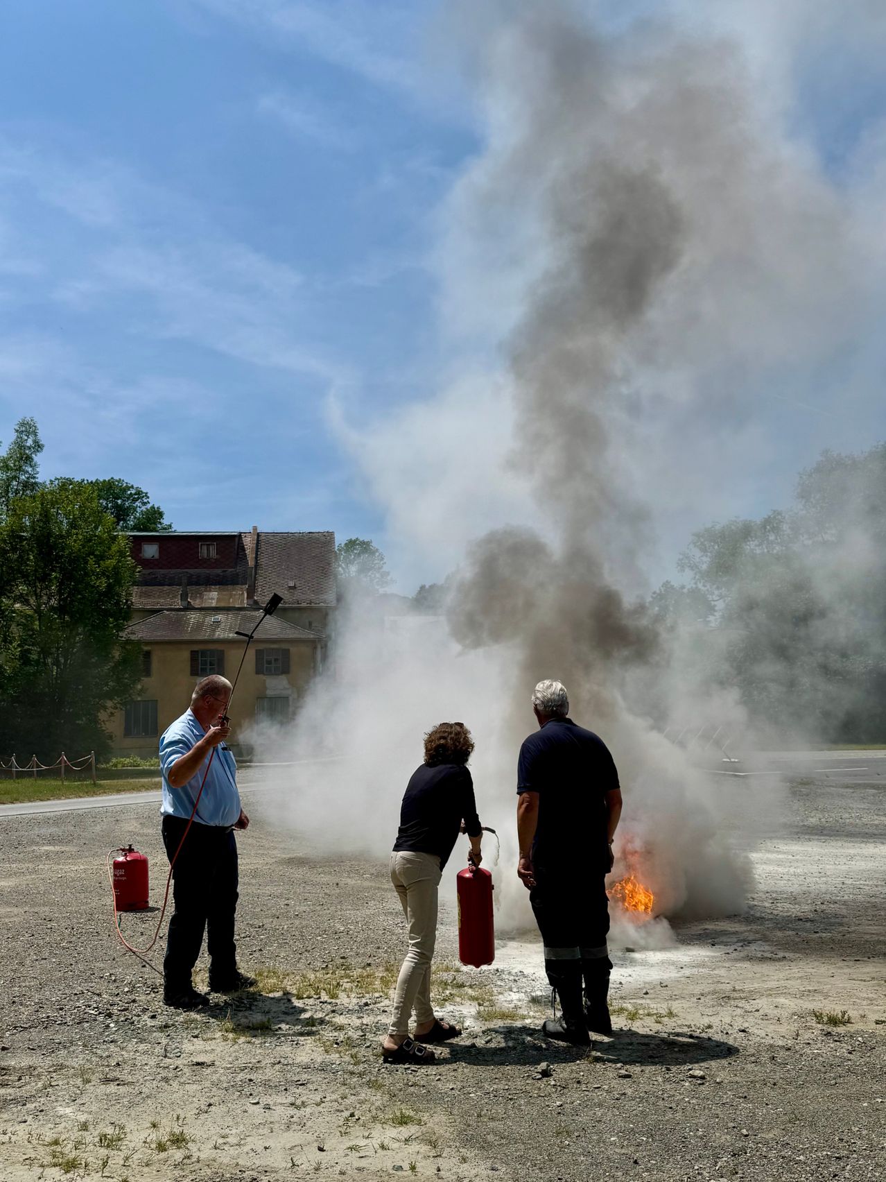 Drei Personen stehen auf einer Straße mit einem Feuerlöscher in der Hand. Einer sprüht Wasser, während die anderen beiden beobachten. Im Hintergrund entsteht ein Feuer, das Rauch abgibt.