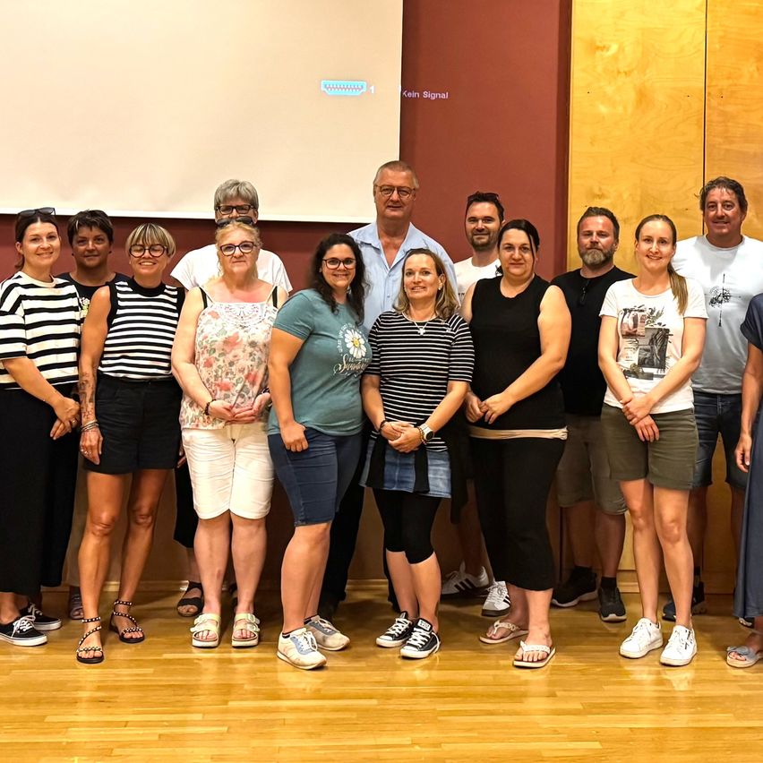 Eine Gruppe von Menschen posiert für ein Foto in einem Raum mit Holzböden und -wänden. Einige tragen Brillen, und eine Projektionsleinwand ist an der Wand montiert.