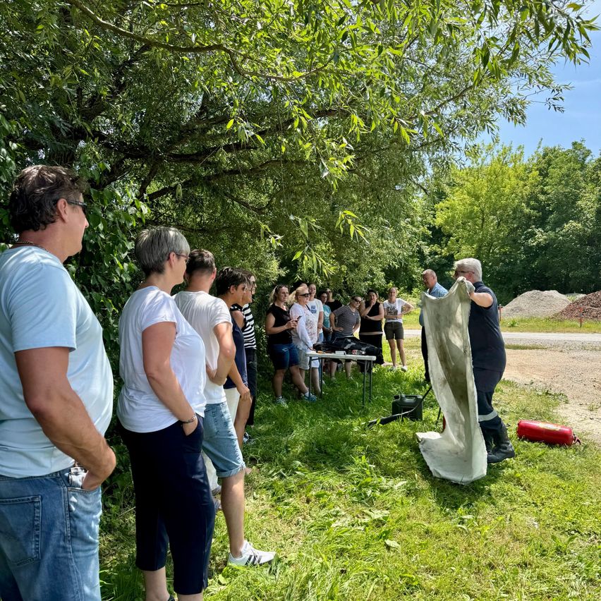 Eine Gruppe von Menschen versammelt sich in einem Grasbereich unter einem Baum, wobei eine Person ein großes weißes Tuch hält. In der Nähe ist ein Tisch mit Gegenständen aufgestellt, und ein Feuerlöscher liegt auf dem Boden. Bäume und eine unbefestigte Straße sind im Hintergrund zu sehen.