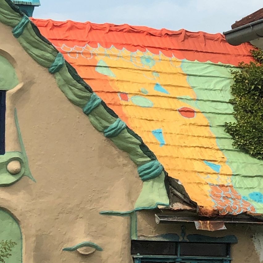 Ein Haus mit einem abstrakten, farbenfrohen und beschädigten Dach. Das Dach zeigt eine Mischung aus Orange, Gelb und Grün. Das Haus hat ein Fenster auf der linken Seite.