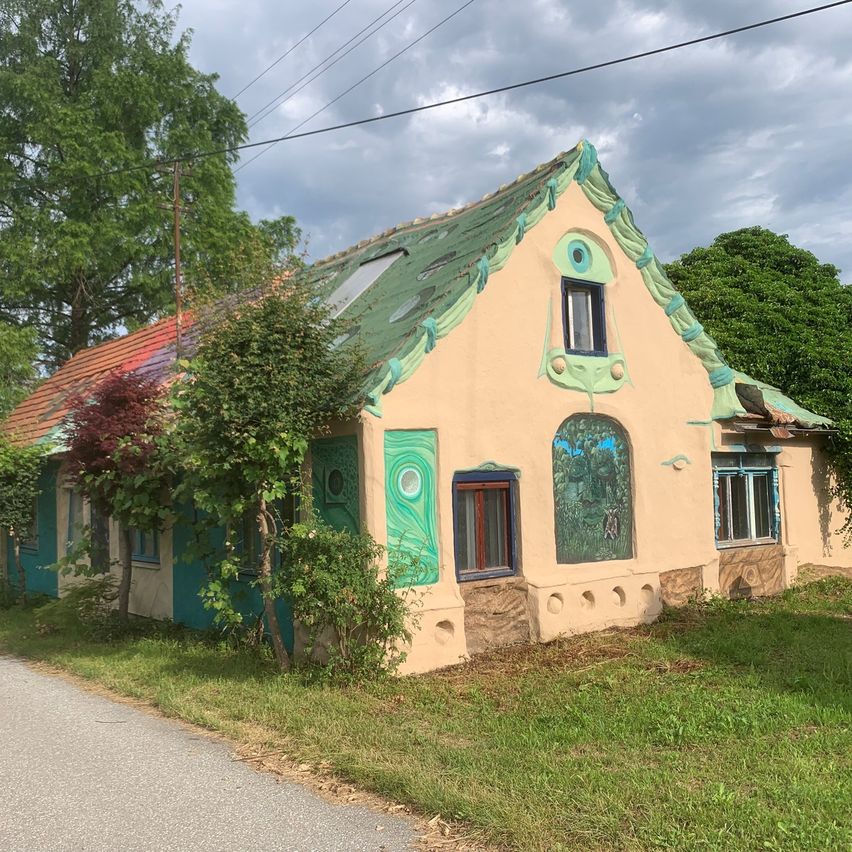 Ein malerisches, künstlerisch bemaltes Haus mit grünem Dach und Solarpanel steht neben einer Straße. Die Außenwände sind mit leuchtenden Wandbildern und aufwendigen Designs verziert.